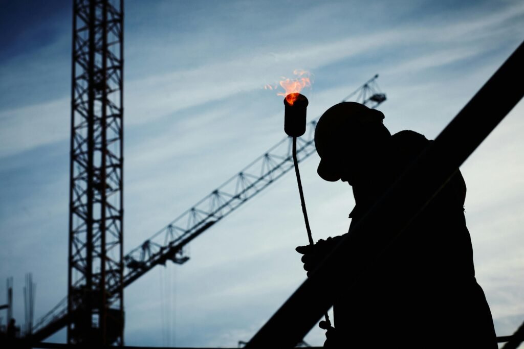 Silhouette of a construction worker using a blowtorch at a building site against a crane-filled skyline.